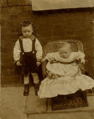 Sepia photograph of a toddler on the left a baby.
					The toddler is wearing bib shorts and prominent boots.
					The baby appears to be in a white dress.