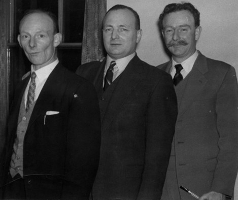 Black and white picture of three men. All are wearing suits and ties.
			The right-most has more hair than the other two - and a moustache.