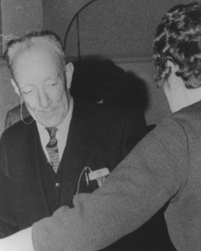 Black and white picture of an older man. Clearly visible is a wired hearing aid.
				In front of him is a back view of a woman with whom he appears to be dancing.